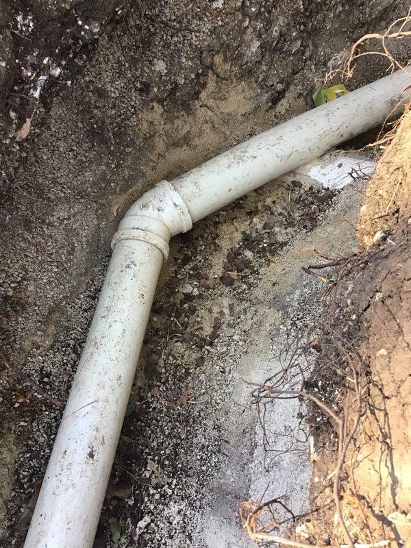 Excavated a trench to expose the underground drain line and completed the drain tunnel work, including installation of PVC drainage piping with a 90-degree elbow fitting and a rubber-banded coupling transition to larger sewer-style pipe. Verified pipe alignment and fitting connections within the trench, with no visible cracking or damage observed on the exposed piping.