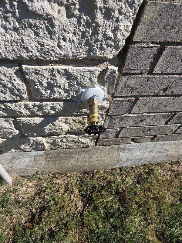 Inspected the front exterior hose bib reported to spray when operated and found the brass sillcock with a cast-iron wheel handle mounted through a limestone and brick wall with a caulked flange. Evaluated the fixture for packing and stem leakage causing the spray condition and documented its condition for repair or replacement of the hose bib.
