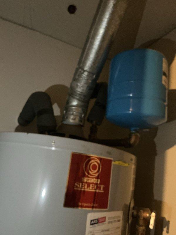 Responded to a garbage disposal complaint and inspected the plumbing system. Documented the existing water heater, including its flue, expansion tank, and insulated supply lines, and photographed the containment basin showing standing moisture and mineral residue at the base of the standpipe. Recorded system conditions to establish a baseline for the account and noted the flue condition for follow-up evaluation.