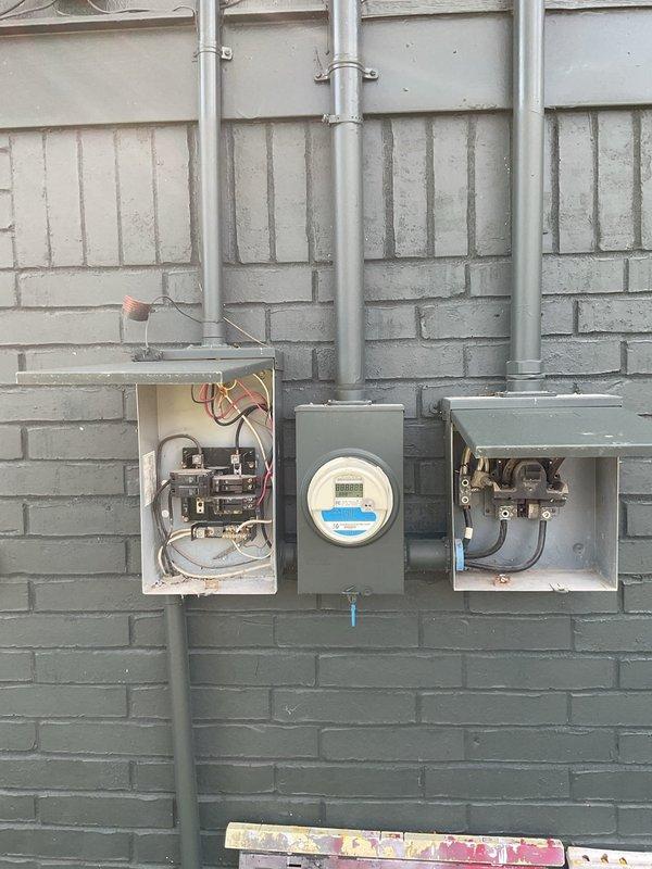 Performed an electrical maintenance inspection on a 50-year-old residence with three breaker boxes (two exterior, one interior). Opened and visually inspected the exterior panel enclosure, meter enclosure, and disconnect/meter socket assembly, evaluating conduit connections, breaker condition, and internal wiring and terminations. Also inspected the workshop receptacles, noting a power strip and extension cord plugged into the outlet and stored materials with hazard labeling nearby.