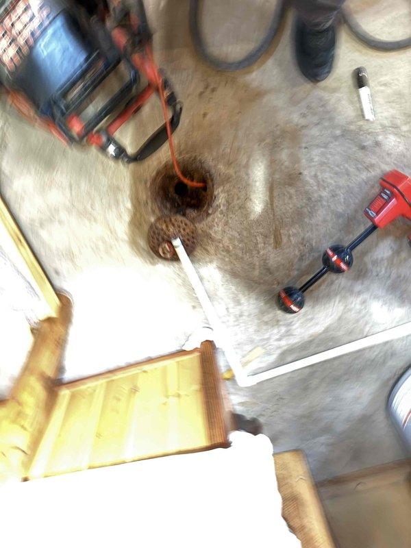 Performed camera inspection of standing drain pipe in basement area to diagnose washing machine drainage backup issue. Accessed the drain system through floor opening, revealing a heavily corroded metal drain cover with significant rust deterioration. Inserted camera equipment to evaluate internal pipe condition and identify potential blockage causing the reported backup problem.