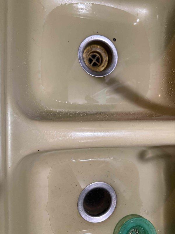 Responded to service call for kitchen sink clogged drain. Inspected P-trap configuration under double basin sink showing significant rust and mineral buildup around drain stoppers. Successfully cleared blockage and restored proper drainage to both basins.