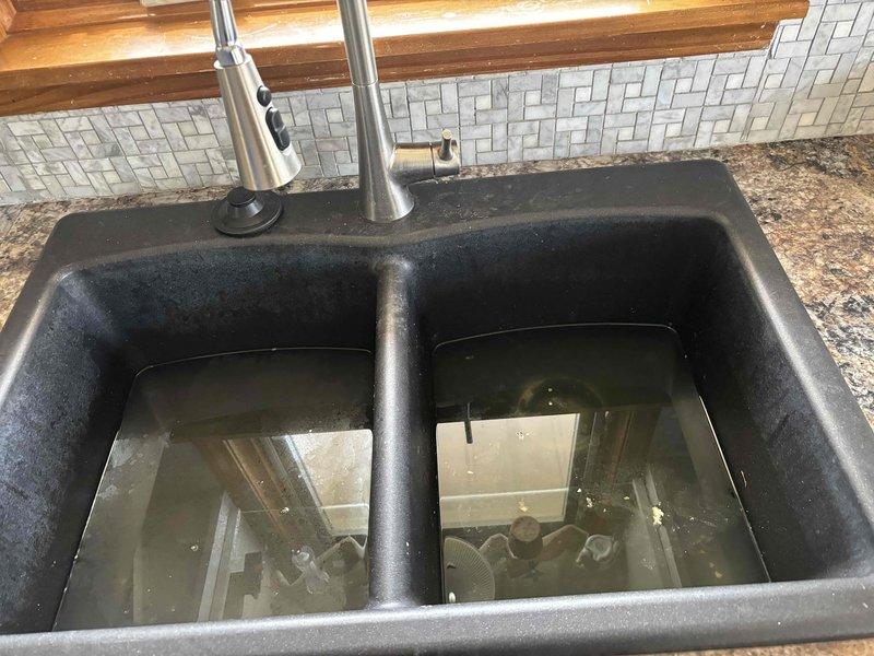 Responded to service call for clogged kitchen sink with standing water in both basins of black composite double-sink. Diagnosed and cleared blockage in the drainage system connected to garbage disposal unit, restoring proper water flow through the P-trap configuration and PVC drain pipes.