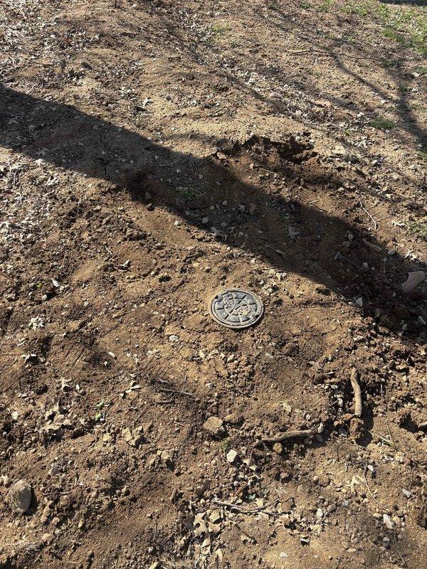 Accessed and serviced lower clean out cap located in yard area. Documented existing ground conditions and clean out accessibility with photographic evidence showing utility access cover embedded in exposed soil with scattered gravel and disturbed earth surrounding the work site.