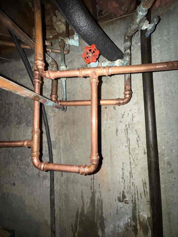 Responded to reported water leak in basement originating from both the main water line and water heater. Upon inspection, discovered severely corroded copper piping with significant oxidation and deterioration throughout the plumbing system. The horizontal water supply line exhibited extensive green and white corrosion buildup, indicating prolonged moisture exposure and advanced pipe degradation. Wall surfaces behind the piping showed vertical streaking patterns consistent with active water seepage. Evaluated the compromised pipe sections, isolation valves, and connection points to identify leak sources. Documented the condition of the aging copper and iron pipe assembly, including deteriorated fittings and joints. The plumbing infrastructure displayed signs of long-term water damage requiring immediate attention to prevent further structural compromise and water waste.