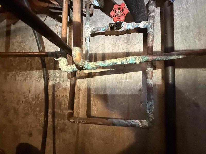 Responded to reported water leak in basement originating from both the main water line and water heater. Upon inspection, discovered severely corroded copper piping with significant oxidation and deterioration throughout the plumbing system. The horizontal water supply line exhibited extensive green and white corrosion buildup, indicating prolonged moisture exposure and advanced pipe degradation. Wall surfaces behind the piping showed vertical streaking patterns consistent with active water seepage. Evaluated the compromised pipe sections, isolation valves, and connection points to identify leak sources. Documented the condition of the aging copper and iron pipe assembly, including deteriorated fittings and joints. The plumbing infrastructure displayed signs of long-term water damage requiring immediate attention to prevent further structural compromise and water waste.