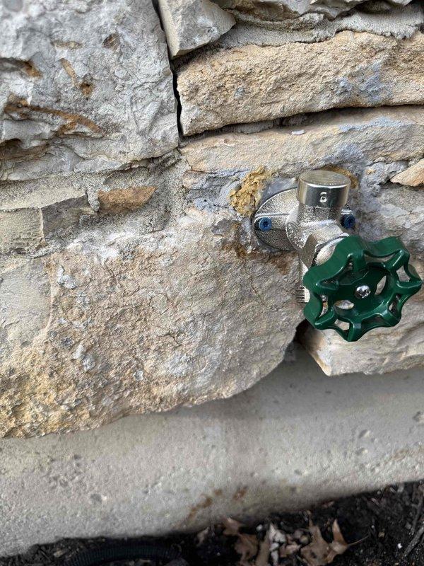 Responded to service call for exterior faucets that were continuously running and failing to shut off properly. Upon arrival, inspected the outside hose bibb assembly with green-handled wheel valve mounted on stone foundation wall. Traced water supply line from exterior valve through wall penetration to access interior plumbing system. Created access opening in interior drywall to expose copper supply piping, wooden framing, and valve connections. Diagnosed faulty valve mechanism preventing proper water shutoff. Repaired/replaced defective components to restore proper operation of exterior faucet control valve. Verified water flow could be fully stopped at valve handle. System tested and confirmed operational with no continued running or leakage.