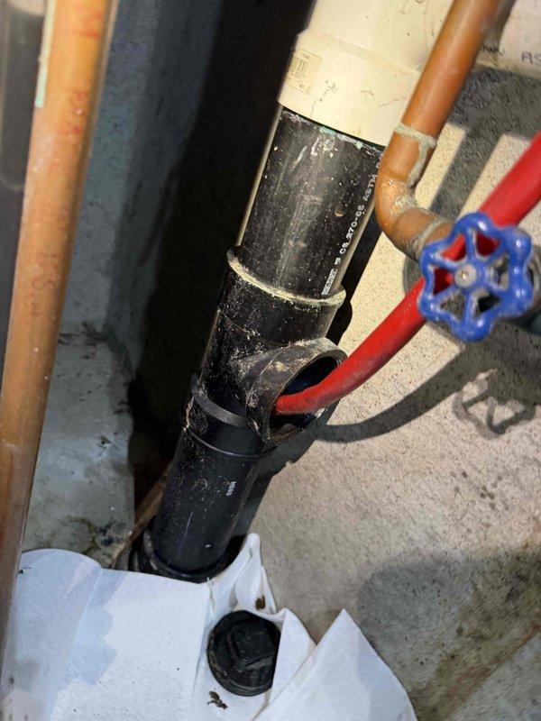 Responded to recall for clogged drain with water backing up from kitchen sink to main bathroom sink. Upon inspection, discovered significant crack in black vertical drain pipe in utility area, which was causing drainage issues and cross-contamination between fixtures. Documented damaged pipe assembly and surrounding plumbing connections including compromised section with irregular opening exposing internal cavity.