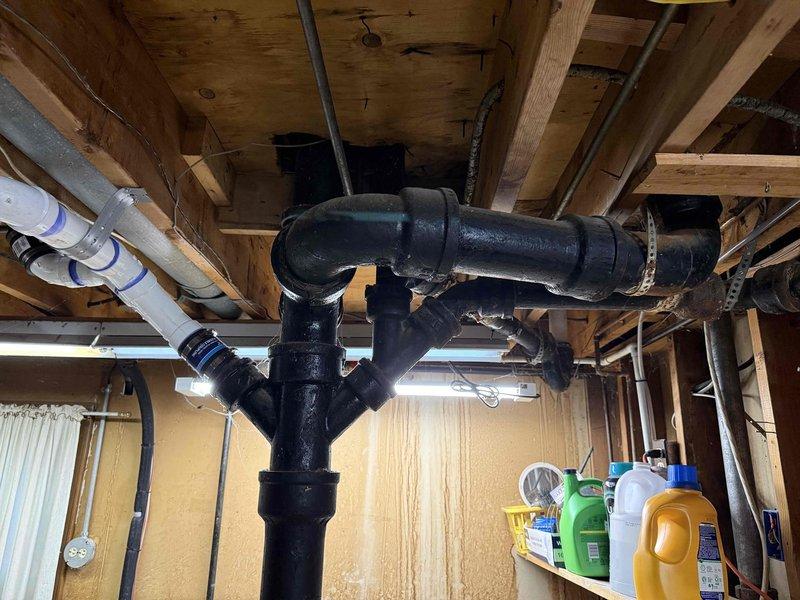 Conducted comprehensive sewer line assessment in basement area to evaluate deteriorating cast iron drain pipes. Inspected exposed black cast iron waste lines running through ceiling joists, documenting multiple cracked sections requiring replacement. Photographed existing plumbing configuration including main vertical stack, horizontal branch lines, and connection points to provide accurate estimate for complete cast iron to PVC conversion.