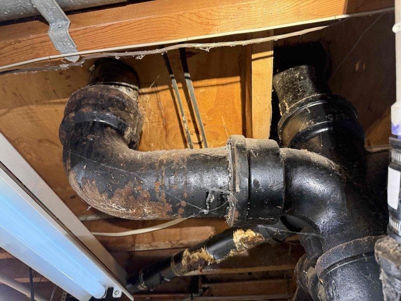 Conducted comprehensive sewer line assessment in basement area to evaluate deteriorating cast iron drain pipes. Inspected exposed black cast iron waste lines running through ceiling joists, documenting multiple cracked sections requiring replacement. Photographed existing plumbing configuration including main vertical stack, horizontal branch lines, and connection points to provide accurate estimate for complete cast iron to PVC conversion.