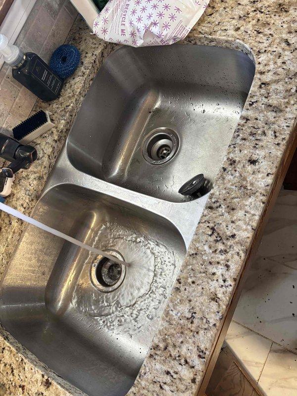 # Plumbing Service - Clogged Drain/Sink

Responded to service call for clogged kitchen drain backing up into dishwasher in early 1960s home with drain located on exterior wall. Upon arrival, inspected double-basin stainless steel sink and under-sink plumbing configuration. Assessed white PVC drain pipes, P-traps, and central drainage system for blockages and proper flow. Cleared obstruction from kitchen drain line, restoring proper drainage and eliminating backup into dishwasher. Verified water flow through both sink basins and confirmed drainage system functioning correctly. Inspected under-sink cabinet area, noting previous moisture exposure on cabinet floor surface. Tested all connections for leaks and proper operation. Additionally addressed secondary issue with toilet not running properly, adjusting fill valve and flush mechanism to restore normal operation. All fixtures tested and confirmed operational upon completion. Documented conditions with photographs of sink area and under