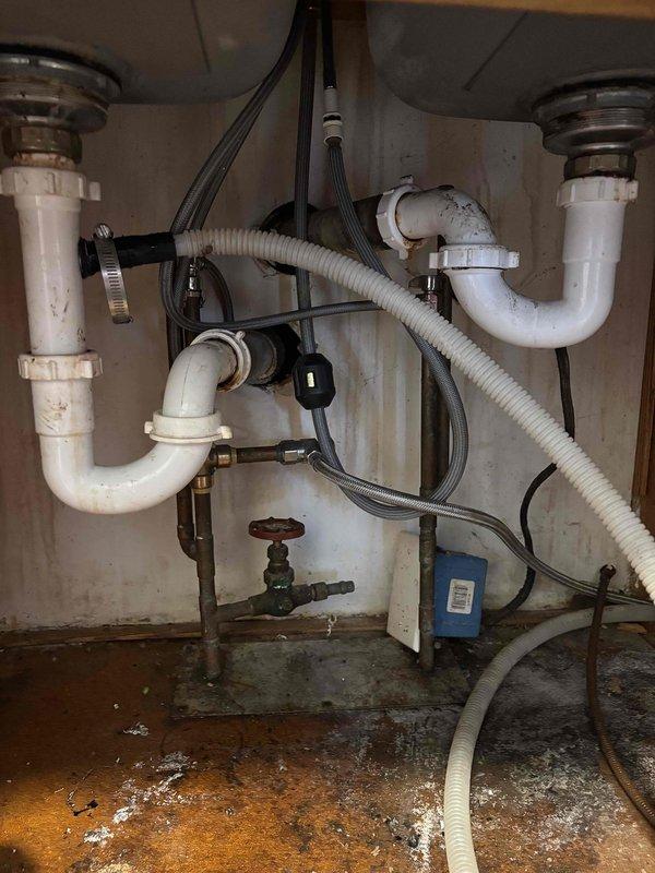 # Plumbing Service - Clogged Drain/Sink

Responded to service call for clogged kitchen drain backing up into dishwasher in early 1960s home with drain located on exterior wall. Upon arrival, inspected double-basin stainless steel sink and under-sink plumbing configuration. Assessed white PVC drain pipes, P-traps, and central drainage system for blockages and proper flow. Cleared obstruction from kitchen drain line, restoring proper drainage and eliminating backup into dishwasher. Verified water flow through both sink basins and confirmed drainage system functioning correctly. Inspected under-sink cabinet area, noting previous moisture exposure on cabinet floor surface. Tested all connections for leaks and proper operation. Additionally addressed secondary issue with toilet not running properly, adjusting fill valve and flush mechanism to restore normal operation. All fixtures tested and confirmed operational upon completion. Documented conditions with photographs of sink area and under