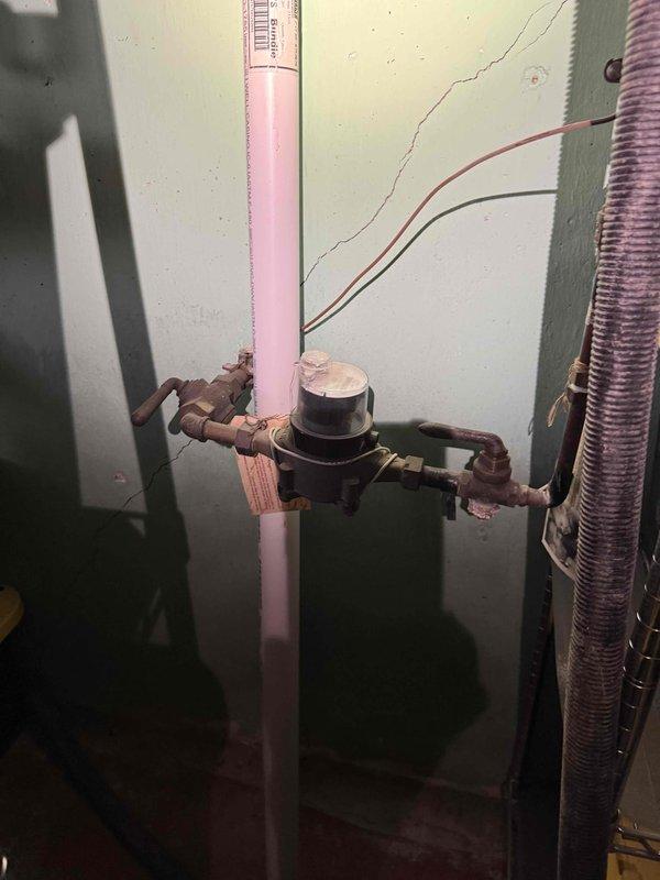 Completed comprehensive whole home plumbing inspection from 8am to 12pm, including tankless water heater flush service. Inspection revealed pink PVC main water line installation in basement utility area with brass shut-off valve assembly showing visible corrosion and weathering on copper fittings requiring monitoring. Documented pressure gauge readings on water system, noting dual-scale gauge displaying measurements in watts and psi with functional black and red indicator needles. All plumbing systems assessed for proper operation, potential leaks, and code compliance throughout the property.