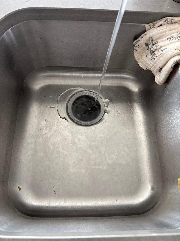 Responded to kitchen sink backup issue occurring during dishwasher operation. Upon inspection, identified significant debris and buildup accumulation in the drain assembly causing restricted water flow. Removed and cleaned the drain strainer, which exhibited heavy grime, tarnishing, and organic matter buildup on the metal cross-strainer mechanism. Cleared the drain line using appropriate tools to restore proper drainage capacity. Tested the system by running the dishwasher to verify water flows freely without backup. Drainage issue resolved successfully.