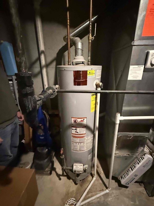Conducted on-site assessment of existing 6-year-old water heater system in basement mechanical room for replacement estimate. Unit currently operational with standard copper supply line connections, exhaust venting, and proximity to expansion tank and HVAC equipment. Documented existing installation configuration including top-mounted pipe connections, metal exhaust ducting, and surrounding utility infrastructure to provide accurate replacement specifications and ensure proper sizing for new water heater installation.
