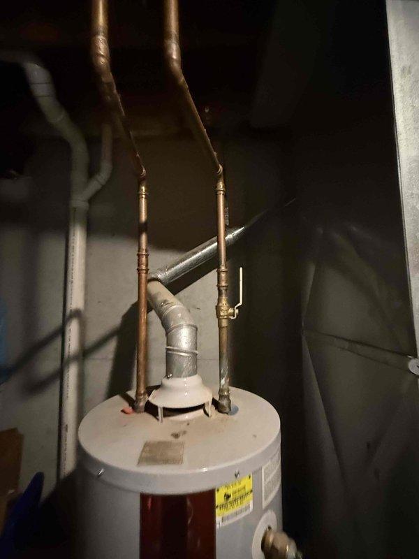 Conducted on-site assessment of existing 6-year-old water heater system in basement mechanical room for replacement estimate. Unit currently operational with standard copper supply line connections, exhaust venting, and proximity to expansion tank and HVAC equipment. Documented existing installation configuration including top-mounted pipe connections, metal exhaust ducting, and surrounding utility infrastructure to provide accurate replacement specifications and ensure proper sizing for new water heater installation.