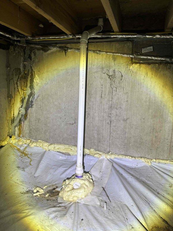Completed plumbing inspection and maintenance visit in basement and bathroom areas. Inspected vertical PVC drainage pipe installation running from ceiling to floor along concrete block wall in crawl space, verifying proper positioning and secure mounting. Examined spray foam insulation application around pipe penetrations and wall perimeter for proper sealing. Assessed moisture barrier installation on floor and noted previous water staining patterns on concrete wall surfaces. In bathroom, inspected toilet installation and accessed confined area behind fixture to examine plumbing connections, including braided stainless steel flexible water supply lines and wall-mounted shut-off valve for proper function and leak-free operation. Documented conditions with photographs for service records.