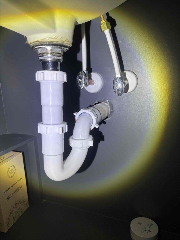 Completed plumbing maintenance inspection under bathroom sink and toilet area. Verified proper installation and functionality of white PVC P-trap assembly with threaded couplings, chrome sink drain basket connection, and dual chrome braided water supply lines with wall-mounted escutcheon plates. All plumbing components showed no signs of leaks, corrosion, or wear, confirming system integrity.