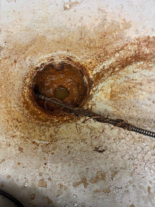 Responded to clogged floor drain in basement. Upon inspection, found heavy rust staining around drain opening with standing water present. Deployed drain snake through the opening to clear blockage. Successfully removed corroded metal spring coil that had become lodged in the drain line, which was the source of the obstruction. Drain now flowing properly with blockage completely cleared.