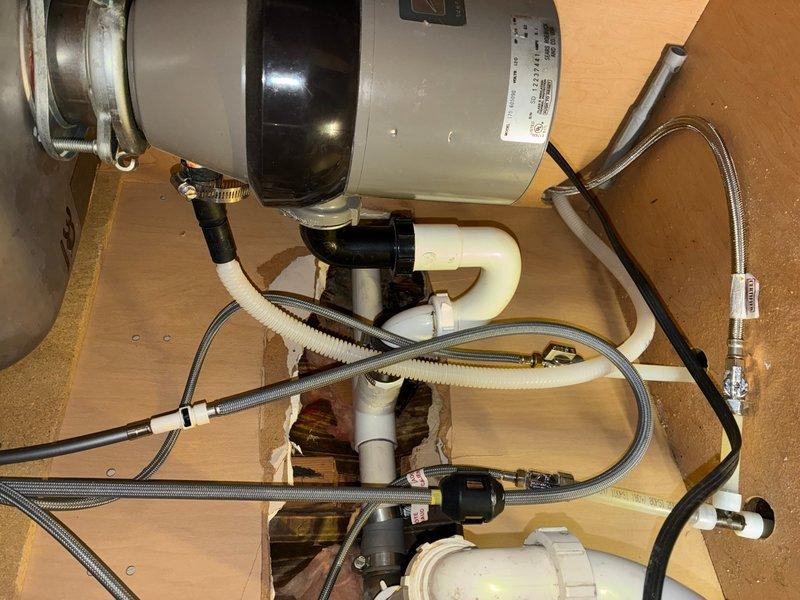 Responded to kitchen plumbing service call regarding improper dishwasher drain line configuration. Upon inspection, identified high loop issue with drain line connection from dishwasher to sink assembly. Corrected dishwasher drain line routing to ensure proper air gap and prevent potential backflow into dishwasher unit. Verified garbage disposal connections, drain trap assembly, and all flexible supply lines were secure and functioning properly. Documented completed installation with photographs showing corrected under-sink plumbing configuration, including disposal unit, PVC drainage pipes, braided stainless steel supply lines, and proper drain line positioning.