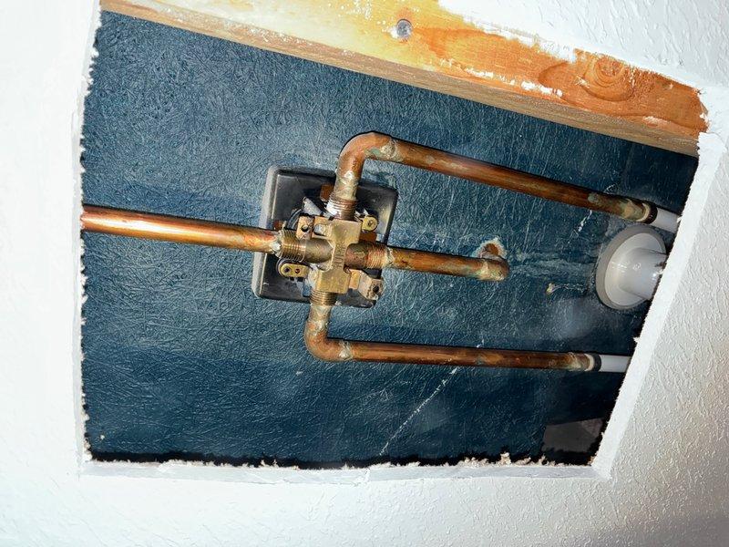Replaced existing shower valve by cutting access opening in sheetrock wall on backside of tub near toilet area to reach plumbing infrastructure. Removed old brass valve assembly with aged copper pipe connections and installed customer-supplied MOEN Caldwell shower valve system. Completed installation of shower trim kit including chrome handheld shower head with flexible braided stainless steel hose and wall mounting bracket on textured gray bathroom wall.
