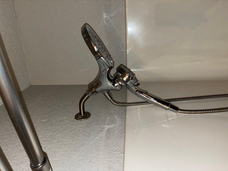 Replaced existing shower valve by cutting access opening in sheetrock wall on backside of tub near toilet area to reach plumbing infrastructure. Removed old brass valve assembly with aged copper pipe connections and installed customer-supplied MOEN Caldwell shower valve system. Completed installation of shower trim kit including chrome handheld shower head with flexible braided stainless steel hose and wall mounting bracket on textured gray bathroom wall.