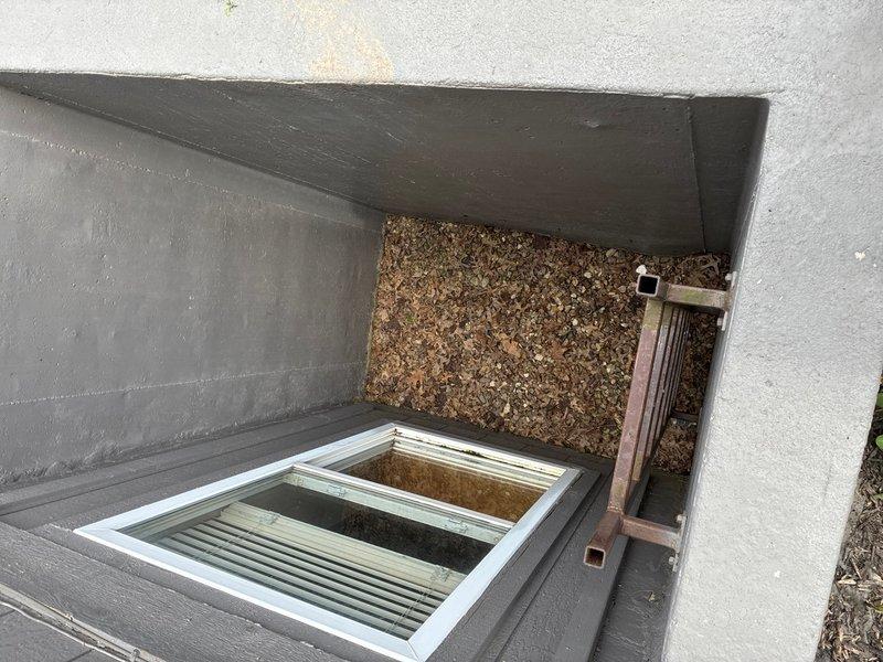 Conducted on-site assessment of basement for sump pump installation in structure currently lacking water management system. Evaluated two existing egress window wells with concrete construction, noting exposed earth and gravel flooring with evidence of organic debris accumulation. Documented current conditions including window well dimensions, drainage patterns, and basement access points to determine optimal sump pump placement and discharge routing for effective groundwater management and flood prevention.