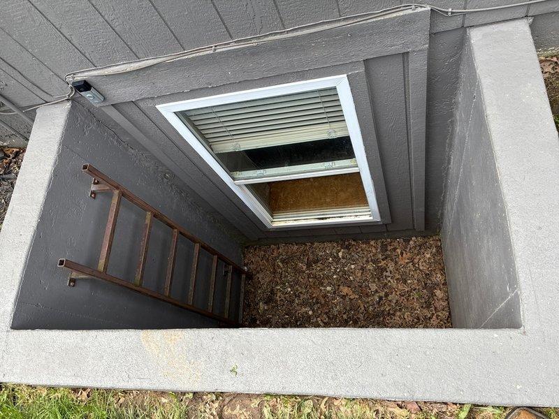 Conducted on-site assessment of basement for sump pump installation in structure currently lacking water management system. Evaluated two existing egress window wells with concrete construction, noting exposed earth and gravel flooring with evidence of organic debris accumulation. Documented current conditions including window well dimensions, drainage patterns, and basement access points to determine optimal sump pump placement and discharge routing for effective groundwater management and flood prevention.