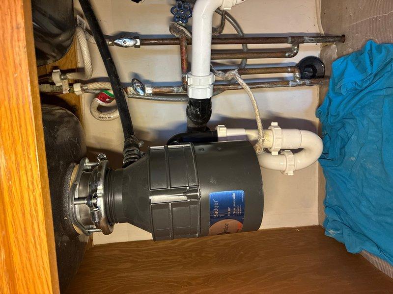 Responded to service call for garbage disposal replacement and leaking pipe investigation in a 40-year-old residence. Upon inspection of the under-sink plumbing system, assessed the existing garbage disposal unit and connected drainage piping for signs of failure and water intrusion. Identified compromised connections and aging drain lines exhibiting corrosion and deterioration at multiple junction points.

Removed the failed garbage disposal unit and disconnected associated drain fittings. Inspected the sink flange, mounting assembly, and discharge elbow for proper sealing and structural integrity. Evaluated the existing PVC drainage configuration, noting darkened areas around joints indicating previous moisture exposure and potential seal failure.

Installed new garbage disposal unit with fresh mounting hardware and properly torqued all connection points to manufacturer specifications. Replaced deteriorated drain fittings and tailpiece connections to ensure watertight seals throughou