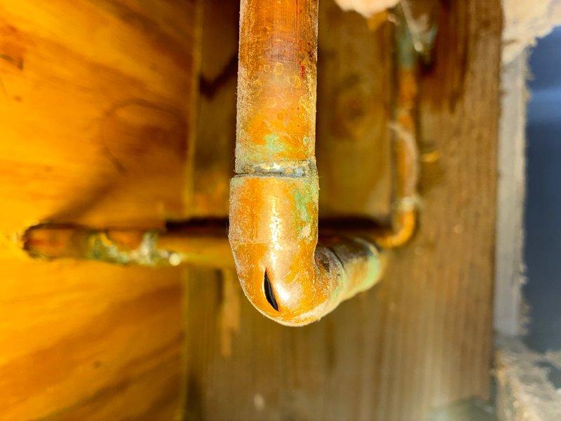 Performed leak location service by cutting access opening in ceiling to expose plumbing system. Investigation revealed corroded copper supply lines with significant oxidation and patina buildup on vertical and horizontal pipe sections, indicating active water intrusion. Access point created through drywall exposed wooden floor joists and fiberglass insulation, allowing for visual inspection and identification of compromised piping requiring repair.