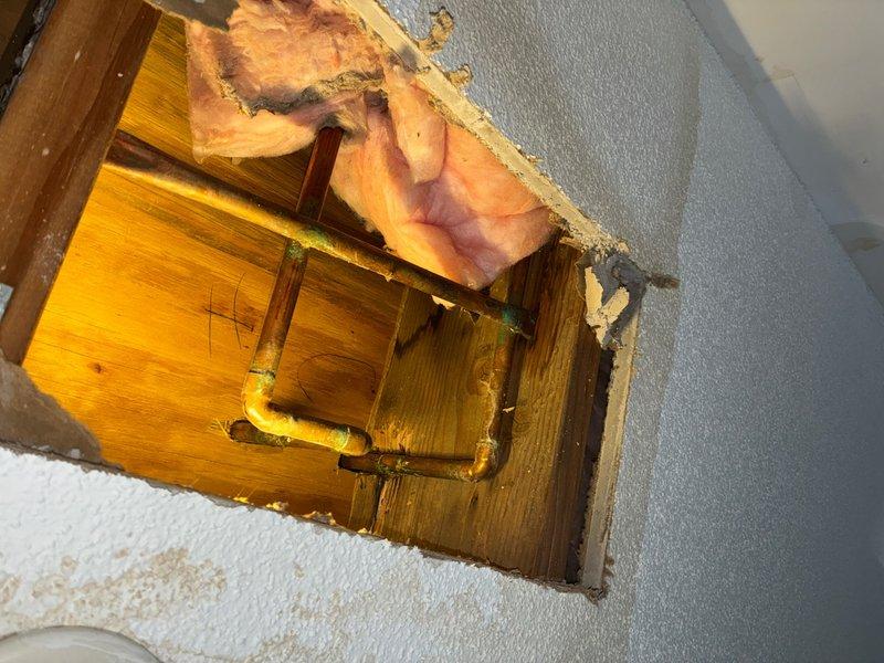 Performed leak location service by cutting access opening in ceiling to expose plumbing system. Investigation revealed corroded copper supply lines with significant oxidation and patina buildup on vertical and horizontal pipe sections, indicating active water intrusion. Access point created through drywall exposed wooden floor joists and fiberglass insulation, allowing for visual inspection and identification of compromised piping requiring repair.