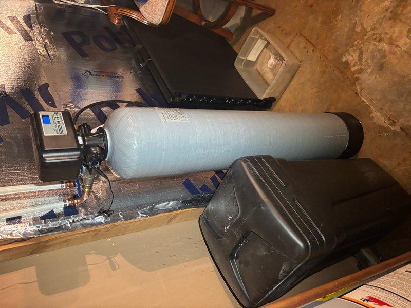 Responded to reported water softener leak in basement. Upon inspection, identified leaking from water softener system connections and piping. Examined copper piping, brass fittings, and control unit connections for source of moisture. Inspected main cylindrical softener tank, brine tank, and associated plumbing components including pressure tank installation in overhead space. Tightened connections at control valve assembly and verified proper sealing of all brass fittings. Checked foam-insulated copper supply lines and expansion tank connections in crawl space above for any secondary leak sources. Tested system operation and monitored for continued leakage. Documented equipment configuration and installation with photographs for service records.