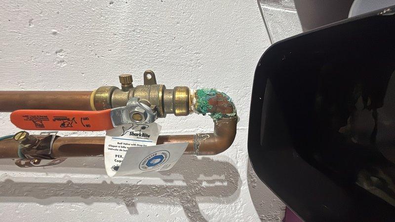 Responded to reported water leaks at main valve line and basement exterior pipe, both exhibiting slow drips with the main line issue occurring seasonally during winter months. Inspected soldered pipe connections at water heater and main shut-off valve assembly, documenting visible corrosion and patina buildup on copper piping and brass fittings. Evaluated kitchen sink low water pressure concern in conjunction with leak assessment. Photographed plumbing configuration including water heater connections, copper supply lines, ball valve assembly, and corroded elbow joint for repair recommendations and further action.