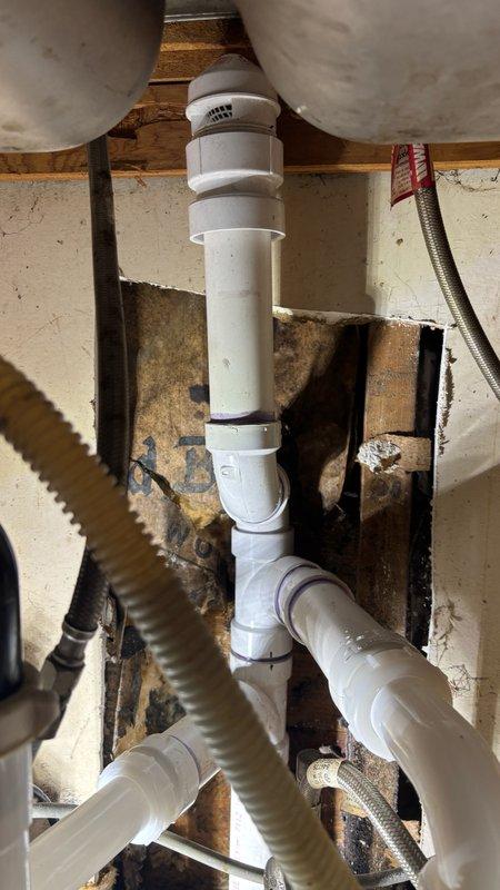 Removed and replaced leaking kitchen drain stack system due to corroded galvanized piping. Removed upper drain line section beneath kitchen cabinet and cut into wall cavity to access drain line tees. Cut out and removed deteriorated 2-inch galvanized pipe from basement level. Installed new PVC drainage piping throughout, reconnected to existing vent system, and repiped connections to double basin sink. Performed complete drainage and leak testing to verify proper functionality of all new installations.