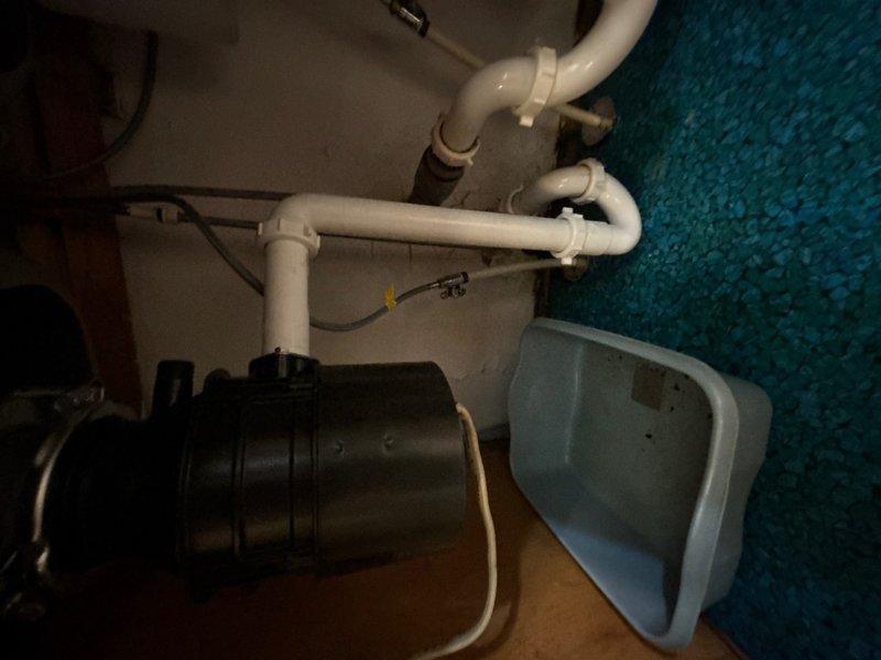 Responded to service call for corroded and fused laundry room water faucets with suspected stem leak preventing proper shutoff, along with leaking garbage disposal in kitchen. Upon inspection, assessed under-sink plumbing configuration including PVC drainage system, trap assembly, and garbage disposal unit connections. Addressed the reported leaking garbage disposal by inspecting mounting assembly, drain connections, and discharge piping for signs of water intrusion. Evaluated corroded washer faucet valves that had become inoperable due to mineral buildup and corrosion, preventing proper water shutoff to the appliance. Documented existing plumbing conditions through photographic evidence showing cabinet interior, drainage components, and disposal unit installation for repair assessment and customer records.