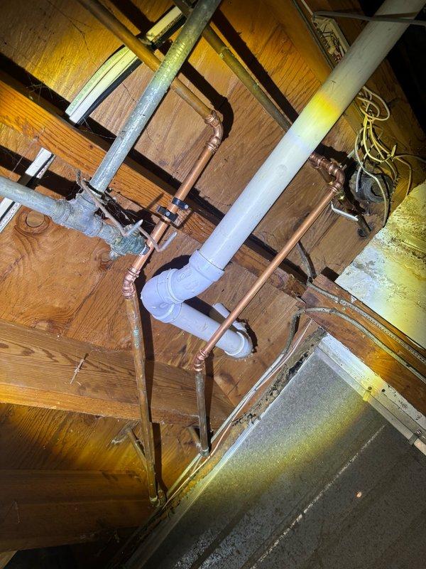 Completed plumbing inspection and documentation of existing basement infrastructure, including copper supply lines, PVC drain piping, and galvanized metal connections running through floor joists. Assessed visible plumbing system components and photographed installation for maintenance records, noting aged copper piping with natural patina and proper junction configurations in crawl space area.