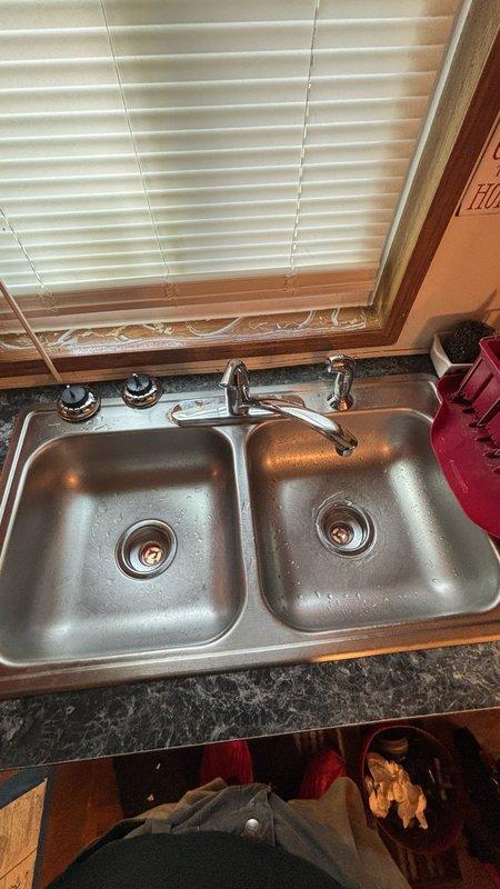 Completed installation of Moen single-handle kitchen faucet with integrated spray function in double-basin stainless steel sink. Installed basket strainer assembly with chrome drain flange and white PVC P-trap configuration. All plumbing connections secured and tested for proper drainage and leak-free operation under cabinet area.