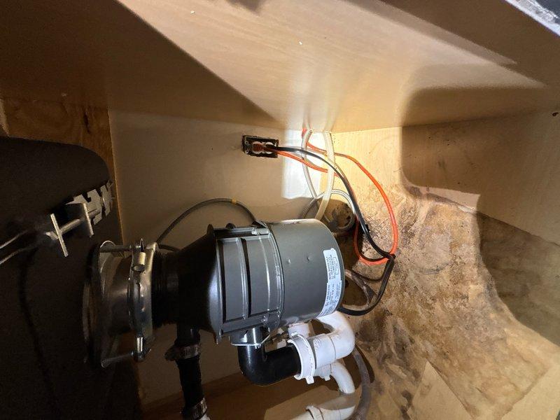 Installed customer-supplied garbage disposal unit under kitchen sink, including complete electrical connections with proper wire terminations and plumbing connections using white PVC drainage pipes and corrugated hoses. Installation includes one-year labor warranty coverage.