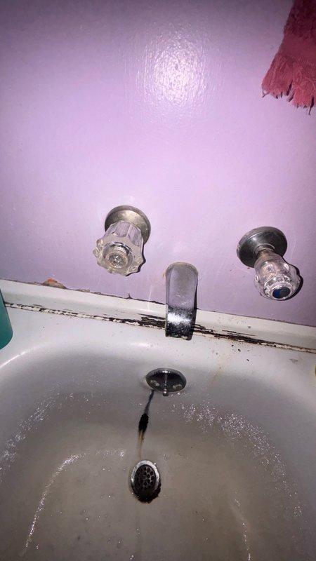 Responded to service call for bathtub faucet replacement in main bathroom. Upon arrival, assessed two wall-mounted chrome faucet fixtures with clear plastic handles showing signs of age and wear. Identified fixtures requiring replacement due to deteriorated condition. Documented existing installation with wall-mounted spout and valve stems protruding from lavender-painted wall surface. Noted visible water staining around drain area and caulking deterioration at wall-to-tub junction indicating potential long-term fixture issues. Prepared estimate for replacement of both bathtub faucet assemblies and recommended addressing caulk seal during installation to prevent future water damage.