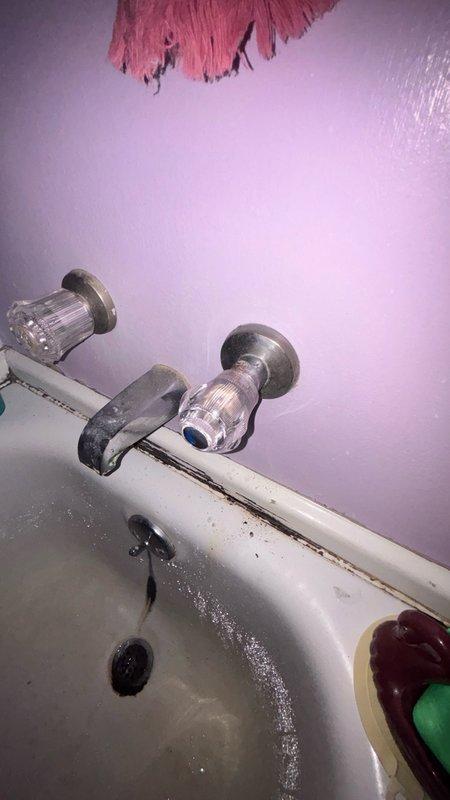 Responded to service call for bathtub faucet replacement in main bathroom. Upon arrival, assessed two wall-mounted chrome faucet fixtures with clear plastic handles showing signs of age and wear. Identified fixtures requiring replacement due to deteriorated condition. Documented existing installation with wall-mounted spout and valve stems protruding from lavender-painted wall surface. Noted visible water staining around drain area and caulking deterioration at wall-to-tub junction indicating potential long-term fixture issues. Prepared estimate for replacement of both bathtub faucet assemblies and recommended addressing caulk seal during installation to prevent future water damage.