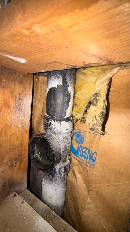 Responded to clogged kitchen drain issue in 40-year-old residential property. Accessed plumbing system through basement crawl space and inspected vertical drain stack. Kitchen sink drainage was completely blocked due to accumulated debris and buildup in the aging black PVC drain line at the Y-junction fitting.

Cleared obstruction using mechanical augering techniques through the cleanout access point. Removed significant grease accumulation and organic matter that had solidified within the pipe over time. Flushed system with water to verify proper flow restoration and confirmed drainage was operating at normal capacity.

Inspected surrounding pipe conditions and noted visible age-related wear on drainage components, including whitish mineral deposits on pipe surfaces. Documented existing moisture damage to adjacent fiberglass insulation and deteriorated wooden structural members around the plumbing chase. Observed advanced decay and dark staining on wall studs indicating long-term wate