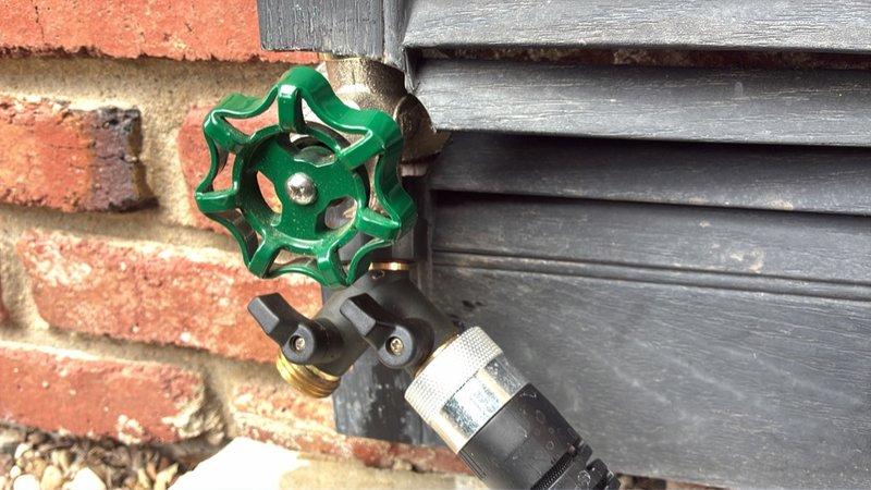 Responded to service call for outdoor faucet assembly experiencing movement and instability issues following installation. Upon inspection, found water spigot with green wheel handle valve, brass valve body, black Y-connector splitter, and quick-connect adapter mounted at brick-to-siding junction. Assessed mounting concerns where vinyl shutter material was inadequate for securing pipe assembly to exterior wall substrate. Evaluated proper anchoring requirements for brick surface to eliminate movement and ensure secure permanent installation of outdoor water supply fixture.