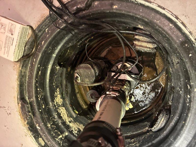 Responded to service call for sump pump inspection in basement of 23-year-old residence. Upon arrival, located and accessed sump pump system installed approximately 2 years prior. Performed comprehensive visual inspection of submersible pump unit, float switch mechanism, discharge piping, electrical connections, and basin condition. Observed pump positioned in standard circular plastic basin with ribbed walls, partially submerged in standing water with murky, greenish-brown appearance indicating normal sediment accumulation. Verified all mechanical components including float switch assembly, electrical wiring, discharge fittings, and clamps were properly secured and functioning. Checked water level and pump operation parameters. Basin and surrounding concrete floor showed typical age-related wear consistent with home's age. No immediate deficiencies, leaks, or operational concerns identified during inspection. System appears to be functioning as designed for groundwater removal and dra