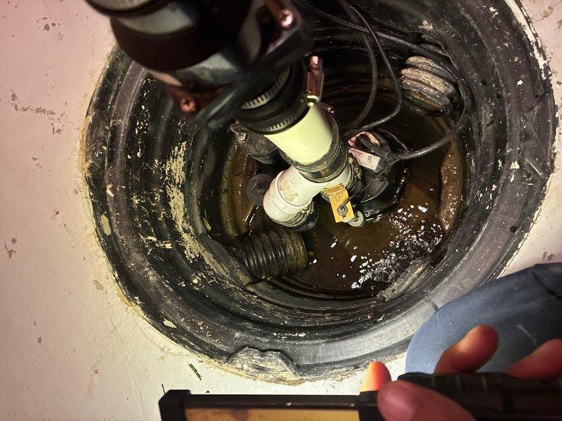Responded to service call for sump pump inspection in basement of 23-year-old residence. Upon arrival, located and accessed sump pump system installed approximately 2 years prior. Performed comprehensive visual inspection of submersible pump unit, float switch mechanism, discharge piping, electrical connections, and basin condition. Observed pump positioned in standard circular plastic basin with ribbed walls, partially submerged in standing water with murky, greenish-brown appearance indicating normal sediment accumulation. Verified all mechanical components including float switch assembly, electrical wiring, discharge fittings, and clamps were properly secured and functioning. Checked water level and pump operation parameters. Basin and surrounding concrete floor showed typical age-related wear consistent with home's age. No immediate deficiencies, leaks, or operational concerns identified during inspection. System appears to be functioning as designed for groundwater removal and dra