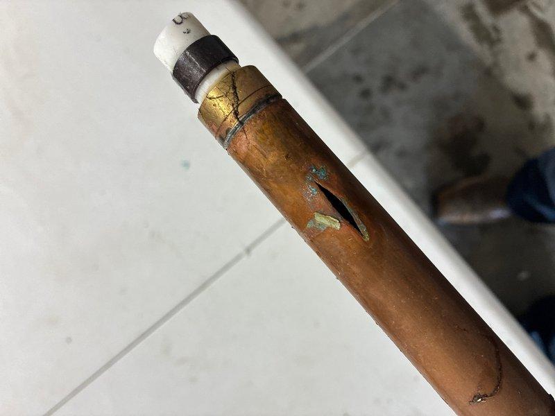 Responded to reported leaking outside faucets at rear of property. Upon inspection, identified severely corroded copper supply pipe with visible horizontal crack and extensive green verdigris oxidation, indicating prolonged water exposure and material failure. Assessed exterior hose bib with green handle wheel mounted on wall above foundation, which was functional but compromised by failed supply line. Documented damaged copper piping showing significant deterioration requiring replacement to restore proper water delivery and prevent continued leakage.
