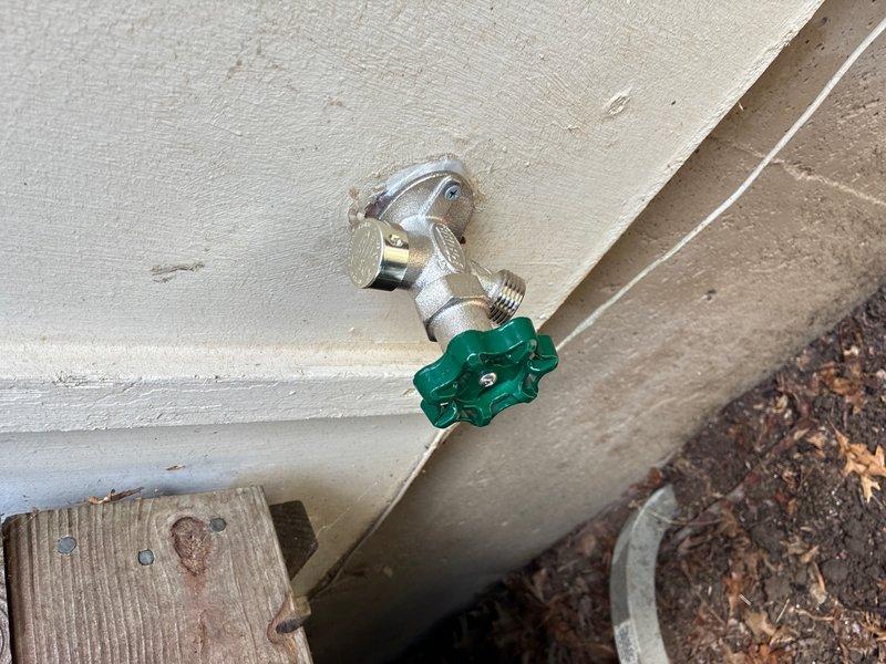 Responded to reported leaking outside faucets at rear of property. Upon inspection, identified severely corroded copper supply pipe with visible horizontal crack and extensive green verdigris oxidation, indicating prolonged water exposure and material failure. Assessed exterior hose bib with green handle wheel mounted on wall above foundation, which was functional but compromised by failed supply line. Documented damaged copper piping showing significant deterioration requiring replacement to restore proper water delivery and prevent continued leakage.