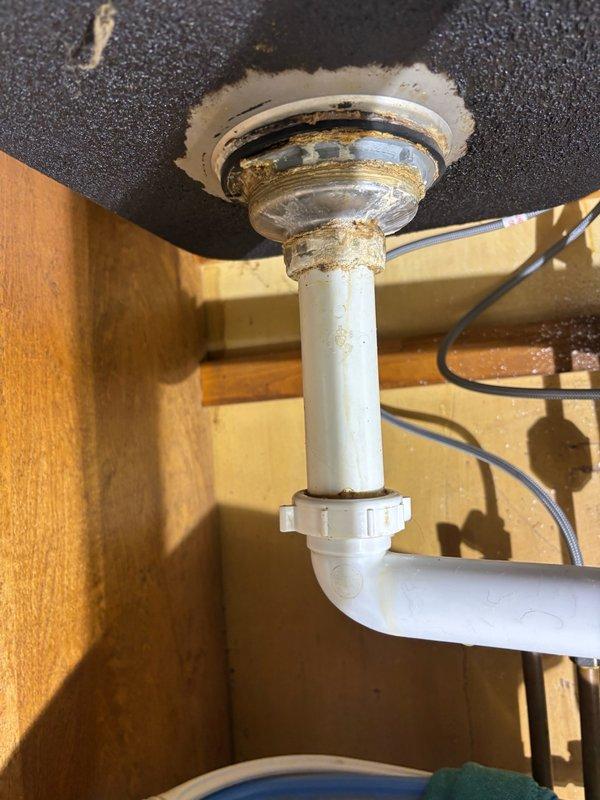 Responded to kitchen sink leak in 50-year-old home. Upon inspection, found deteriorated drain assembly with visible staining and residue buildup around connection points between chrome drain basket and PVC tailpiece. Assessed P-trap configuration and identified leak source at threaded connections where metal components meet plastic piping. Disassembled drain assembly, cleaned all connection surfaces, replaced worn gaskets and seals, and reassembled with proper torque specifications. Verified all connections were secure and tested system under full water flow to confirm leak was eliminated. All plumbing hardware restored to proper working condition with no active leaks detected upon completion.