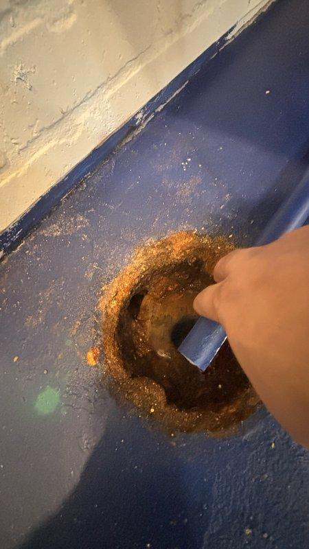 Responded to service call for main line drain cleanout. Upon arrival, accessed the main cleanout point located in floor area showing significant rust and corrosion around the drain opening. Performed mechanical cable cleaning of the main sewer line to remove blockage and restore proper drainage flow. Inspected underside plumbing configuration, noting existing P-trap assembly with white PVC connections to older cast iron piping system. Observed heavy rust staining and corrosion on surrounding surfaces indicating prolonged moisture exposure. Successfully cleared obstruction from main line and verified drainage restoration. Documented existing conditions including deteriorated floor surface and corroded plumbing components for service records.
