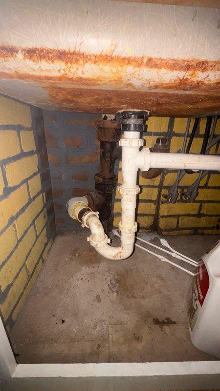Responded to service call for main line drain cleanout. Upon arrival, accessed the main cleanout point located in floor area showing significant rust and corrosion around the drain opening. Performed mechanical cable cleaning of the main sewer line to remove blockage and restore proper drainage flow. Inspected underside plumbing configuration, noting existing P-trap assembly with white PVC connections to older cast iron piping system. Observed heavy rust staining and corrosion on surrounding surfaces indicating prolonged moisture exposure. Successfully cleared obstruction from main line and verified drainage restoration. Documented existing conditions including deteriorated floor surface and corroded plumbing components for service records.