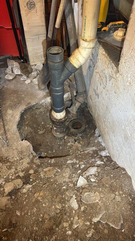 Conducted comprehensive on-site assessment of basement sewer and drain line system to provide estimate for necessary repairs and replacements. Inspection revealed severely deteriorated concrete flooring around main sewer line penetrations, with extensive fragmentation and soil exposure creating potential structural and sanitary concerns. Evaluated condition of existing PVC drain lines, including vertical stack and angled lateral connections, noting age-related discoloration and wear patterns. Documented compromised floor conditions requiring concrete removal and replacement to properly access and service underground sewer lines. Assessed overhead plumbing infrastructure, water supply lines, and electrical routing to determine scope of work and potential conflicts during excavation and installation phases.